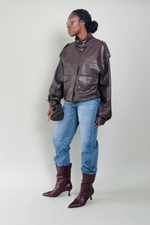 Woman wearing a brown leather jacket, blue jeans, and maroon boots on a plain background