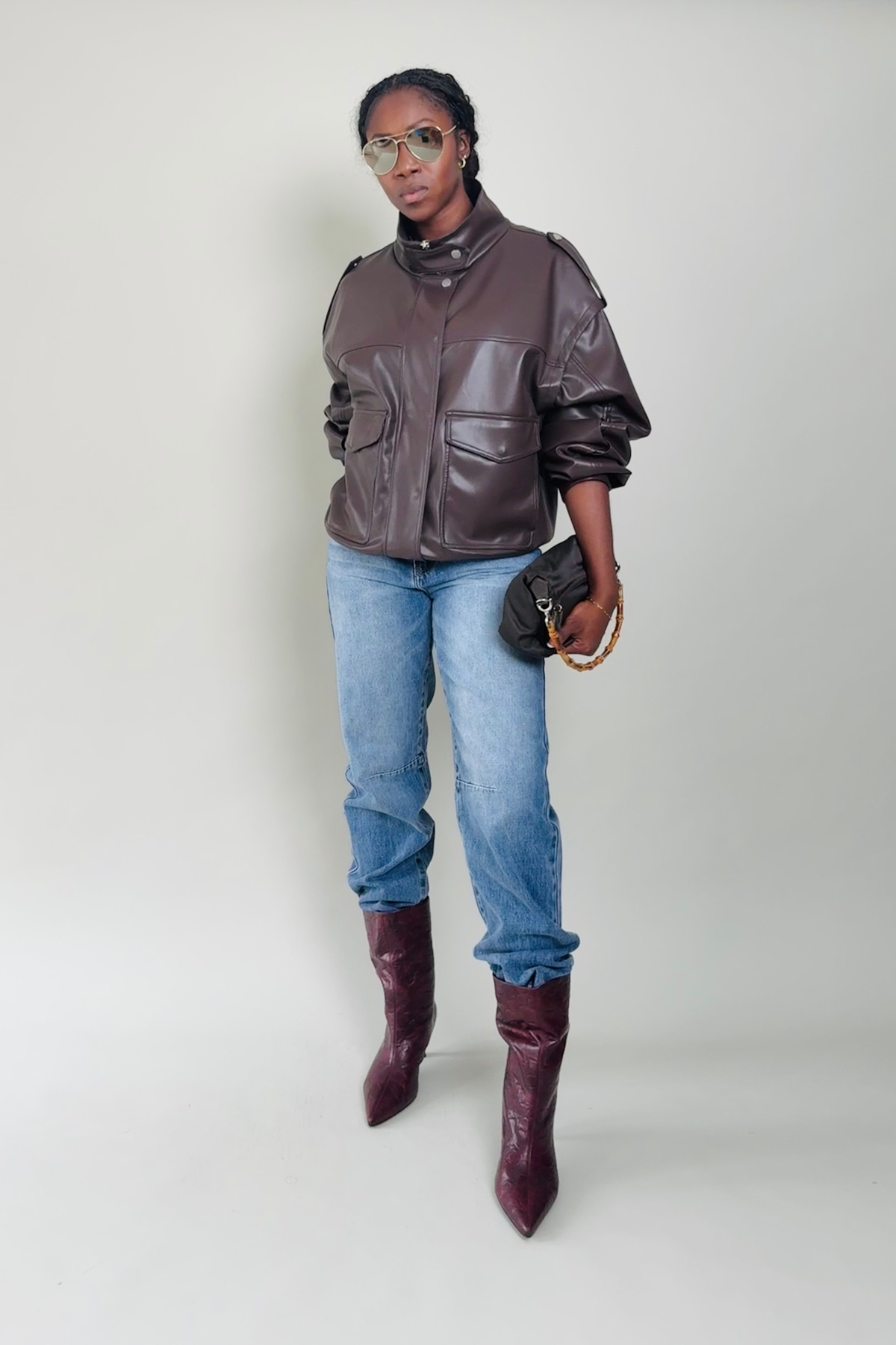 Woman wearing a brown leather jacket, blue jeans, and maroon boots on a plain background