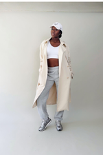 Person wearing a beige trench coat, white top, gray pants, and white sneakers against a plain background