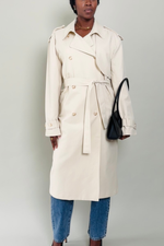 Woman wearing a beige trench coat and blue jeans against a plain background