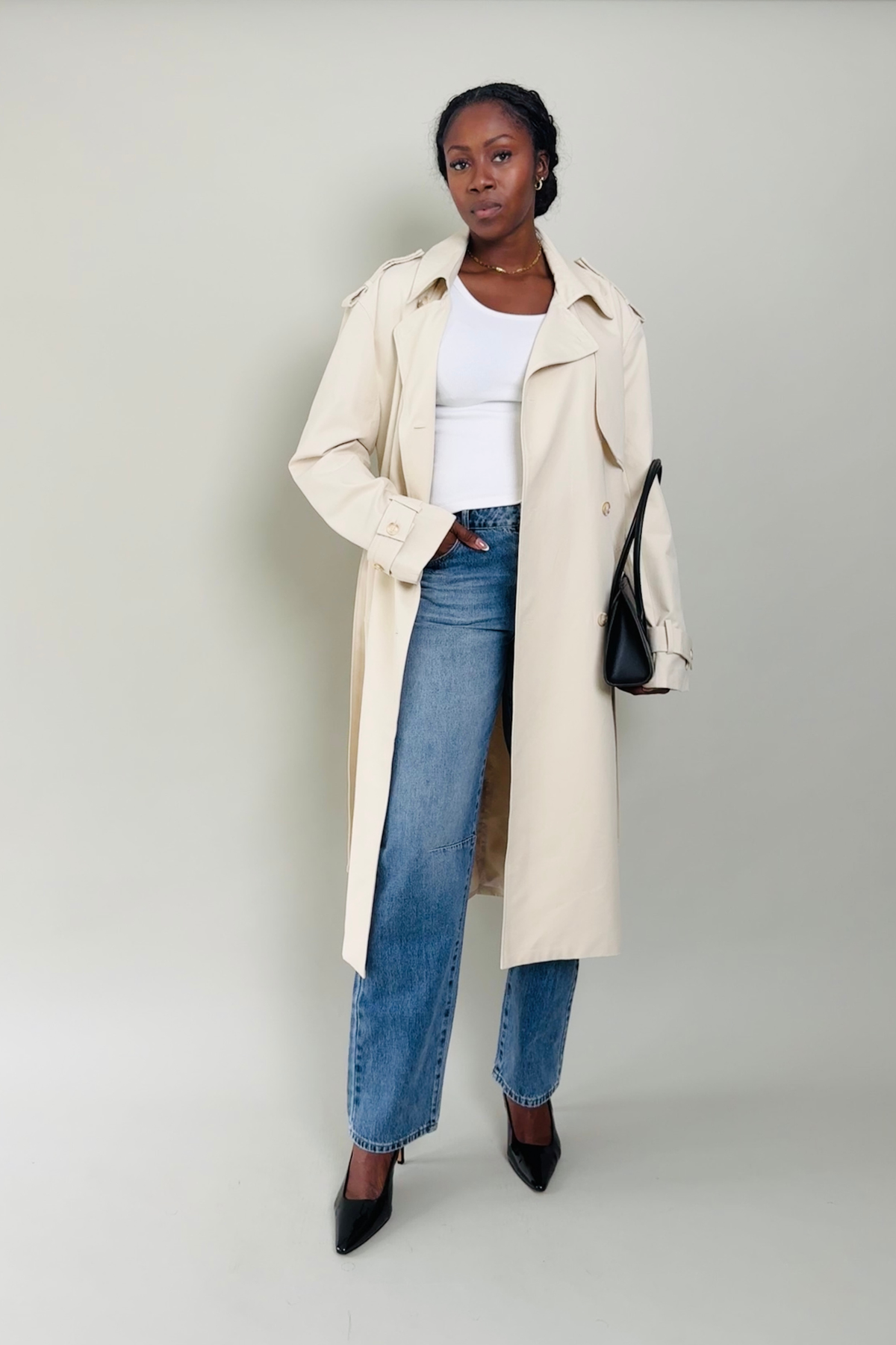 Woman wearing a beige trench coat, white top, blue jeans, and black shoes on a plain background