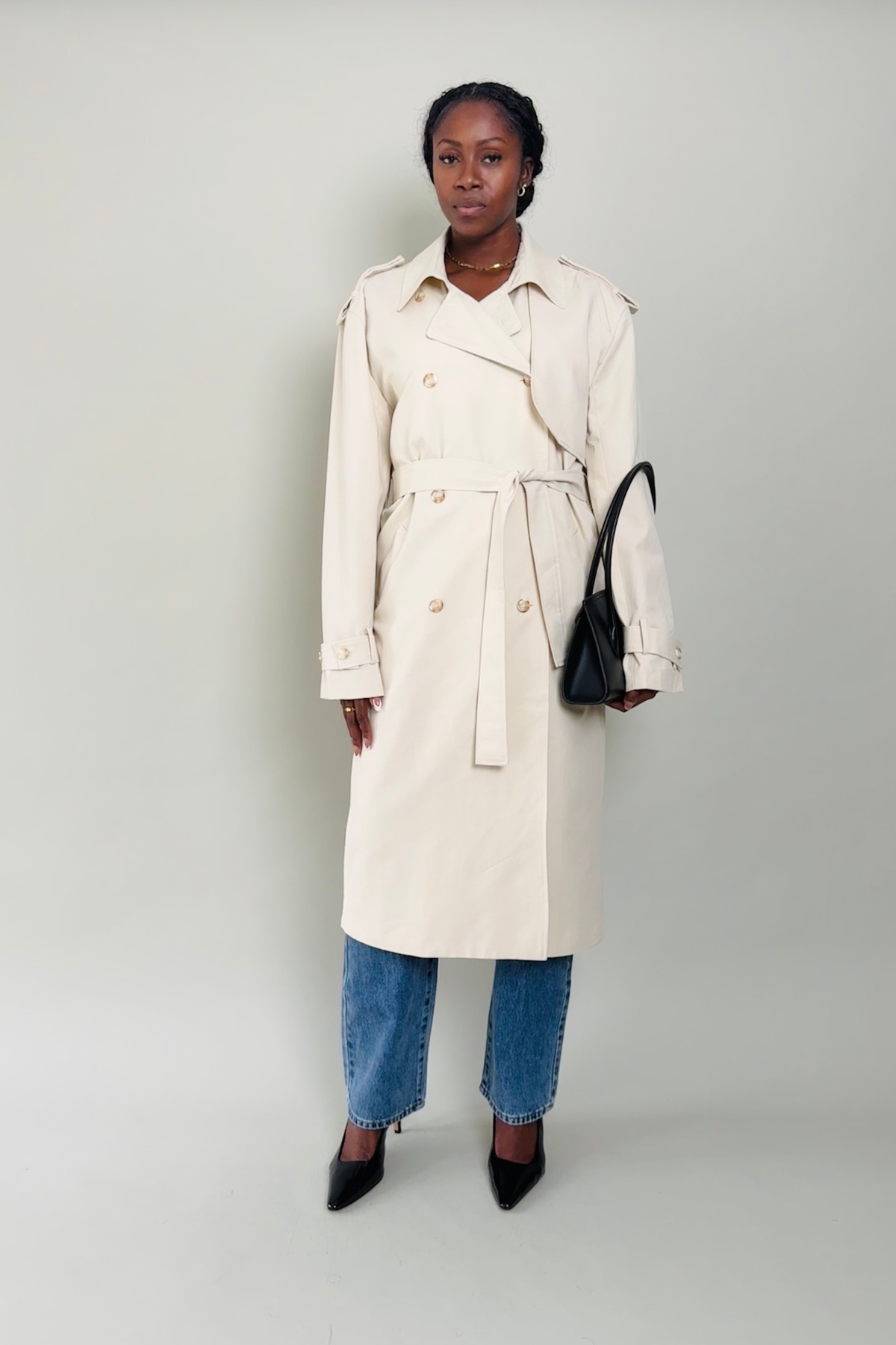 Woman wearing a beige trench coat with blue jeans and black shoes on a plain background