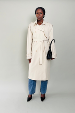 Woman wearing a beige trench coat with blue jeans and black shoes on a plain background
