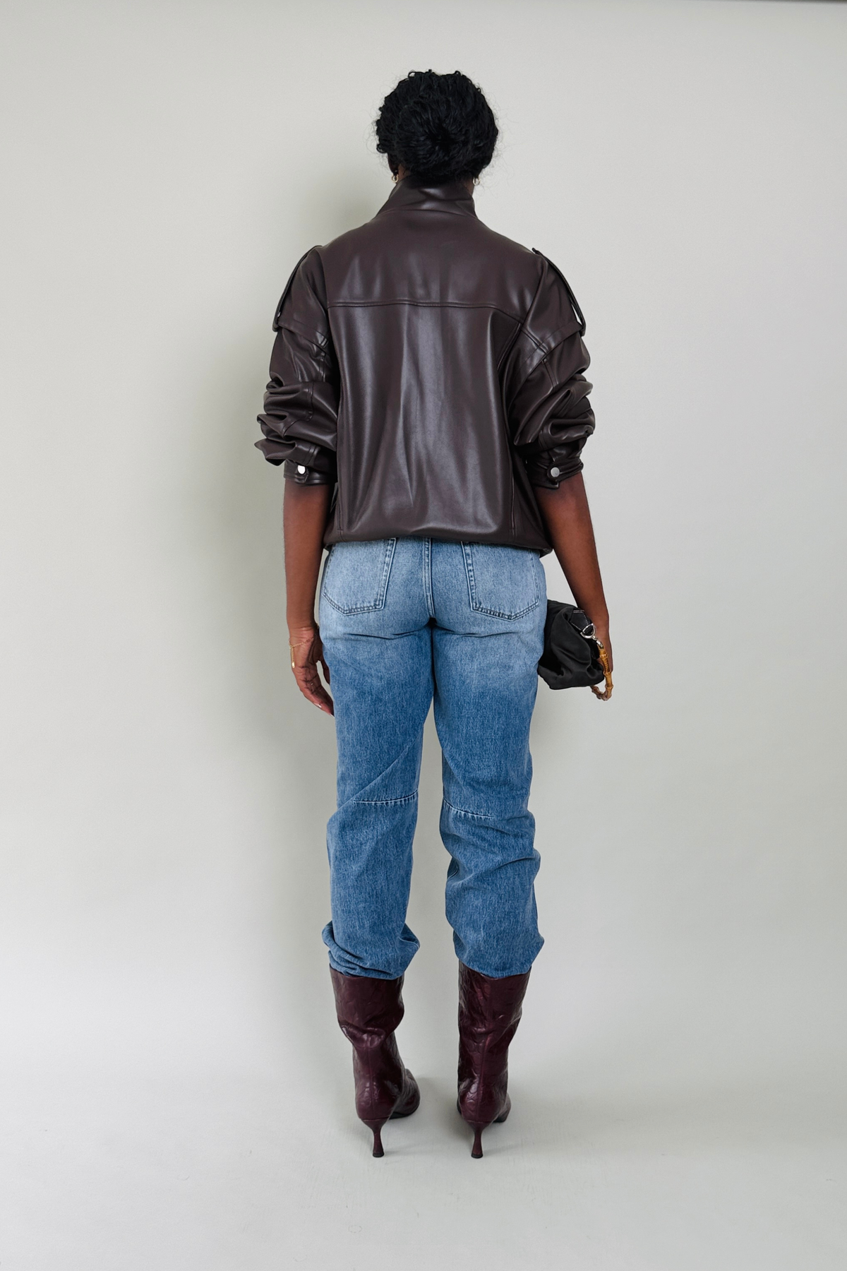 Woman wearing a brown leather jacket, blue jeans, and maroon boots on a plain background