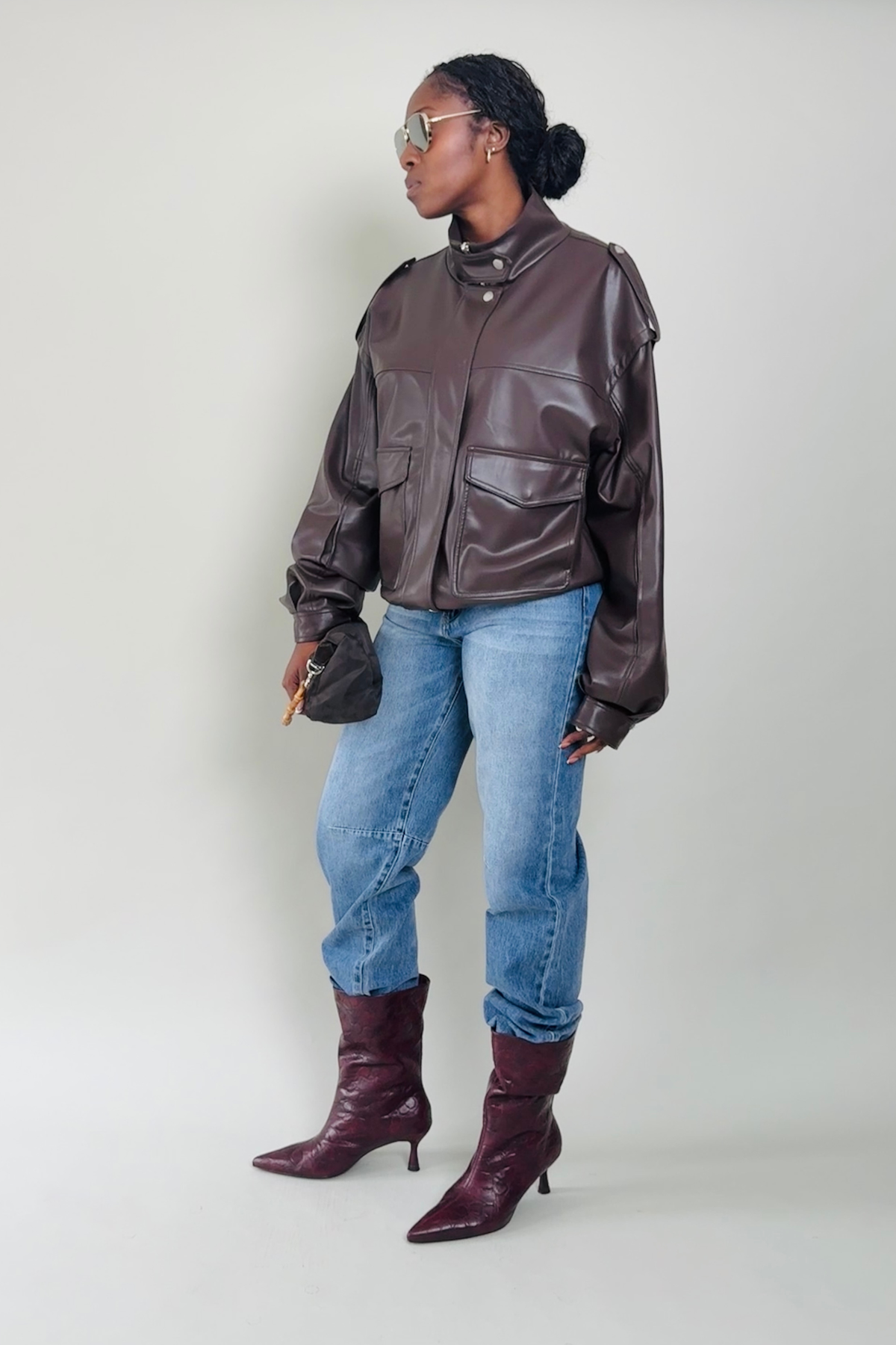 Woman wearing a brown leather jacket, blue jeans, and maroon boots on a plain background
