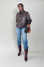Woman wearing a brown leather jacket, blue jeans, and maroon boots on a plain background