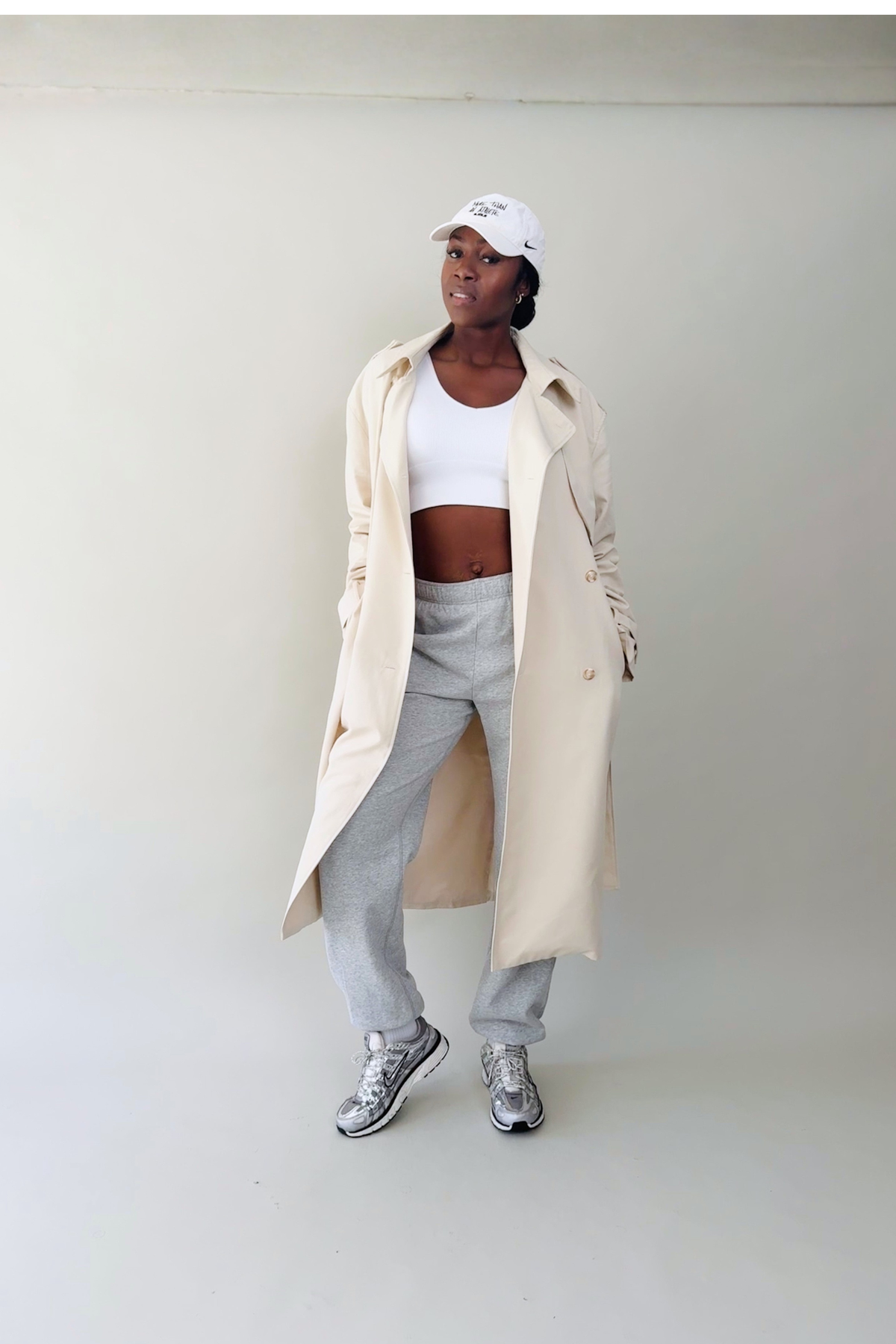 Person wearing a beige trench coat, white top, gray pants, and white sneakers against a plain background