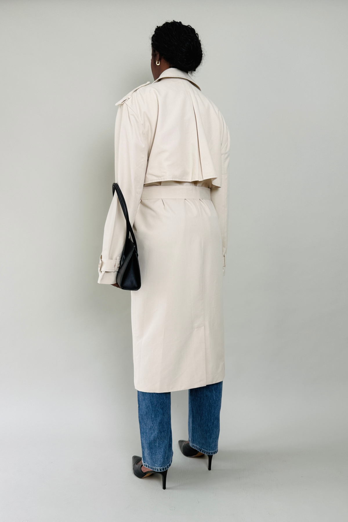 Woman wearing a beige trench coat with blue jeans and black shoes on a plain background
