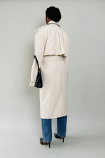 Woman wearing a beige trench coat with blue jeans and black shoes on a plain background